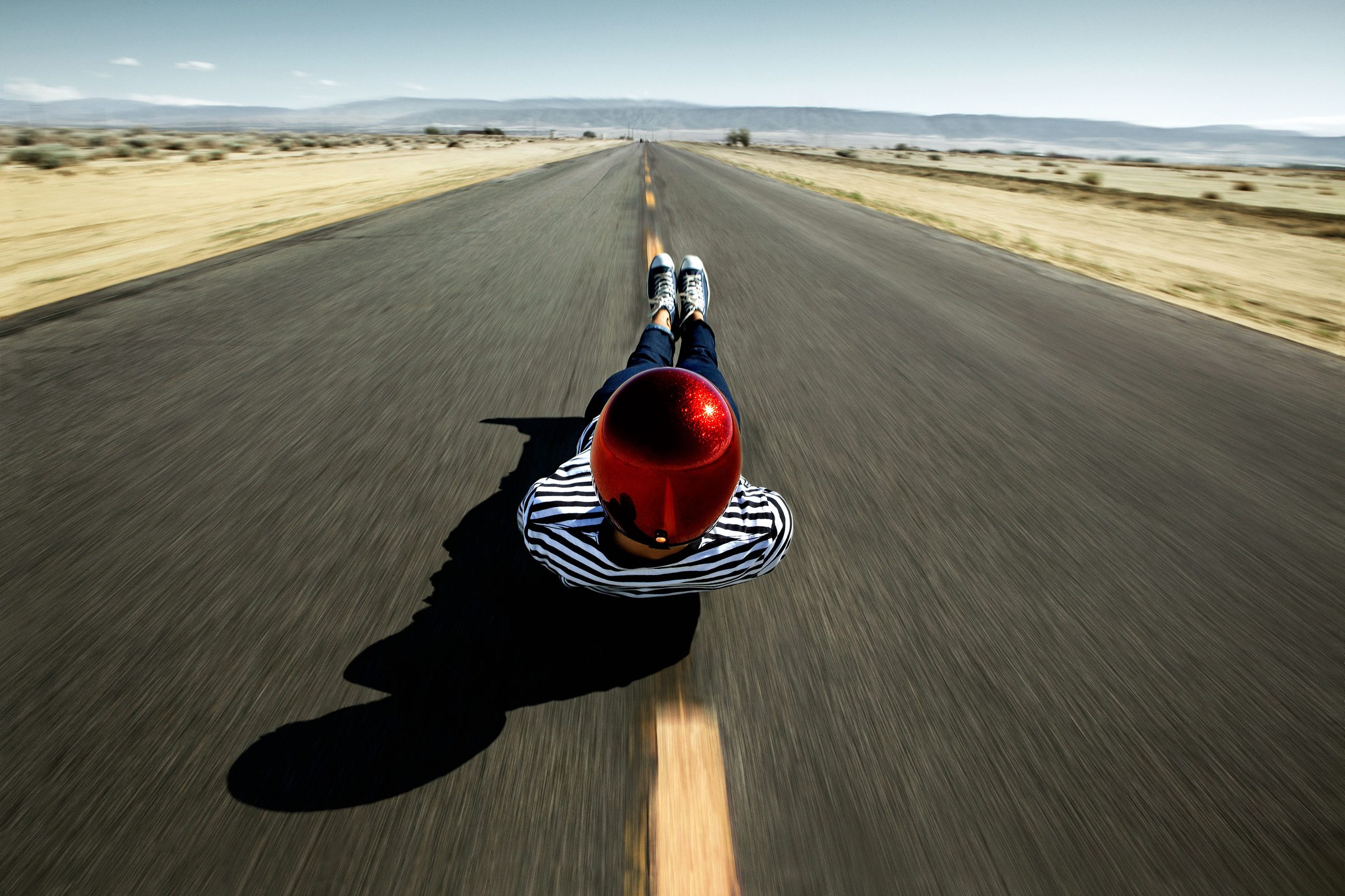 A longboarder in a red helmet and striped tee bombs the centerline of an empty desert highway.