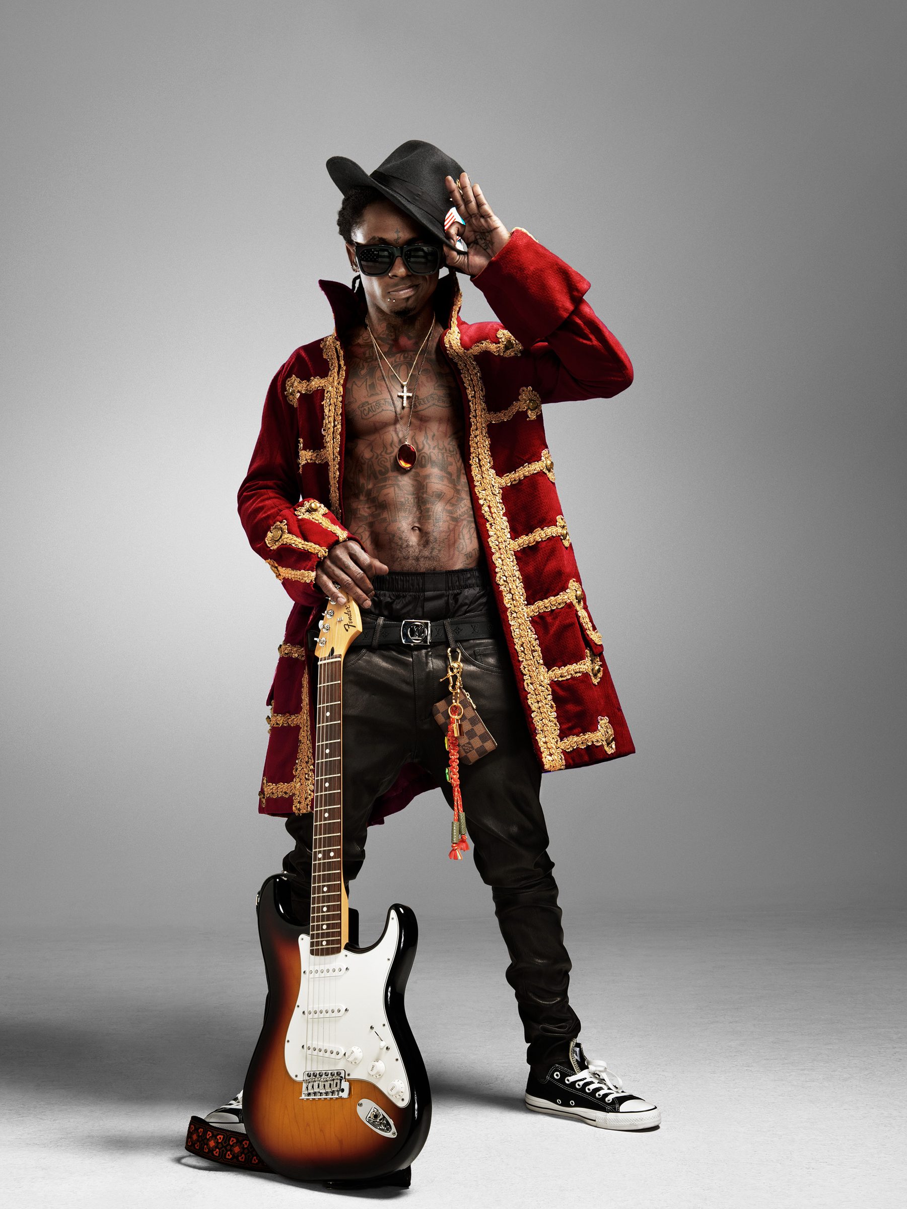 Full-length: crimson military coat, black hat, and a sunburst Stratocaster at his side as Lil Wayne throws a salute.