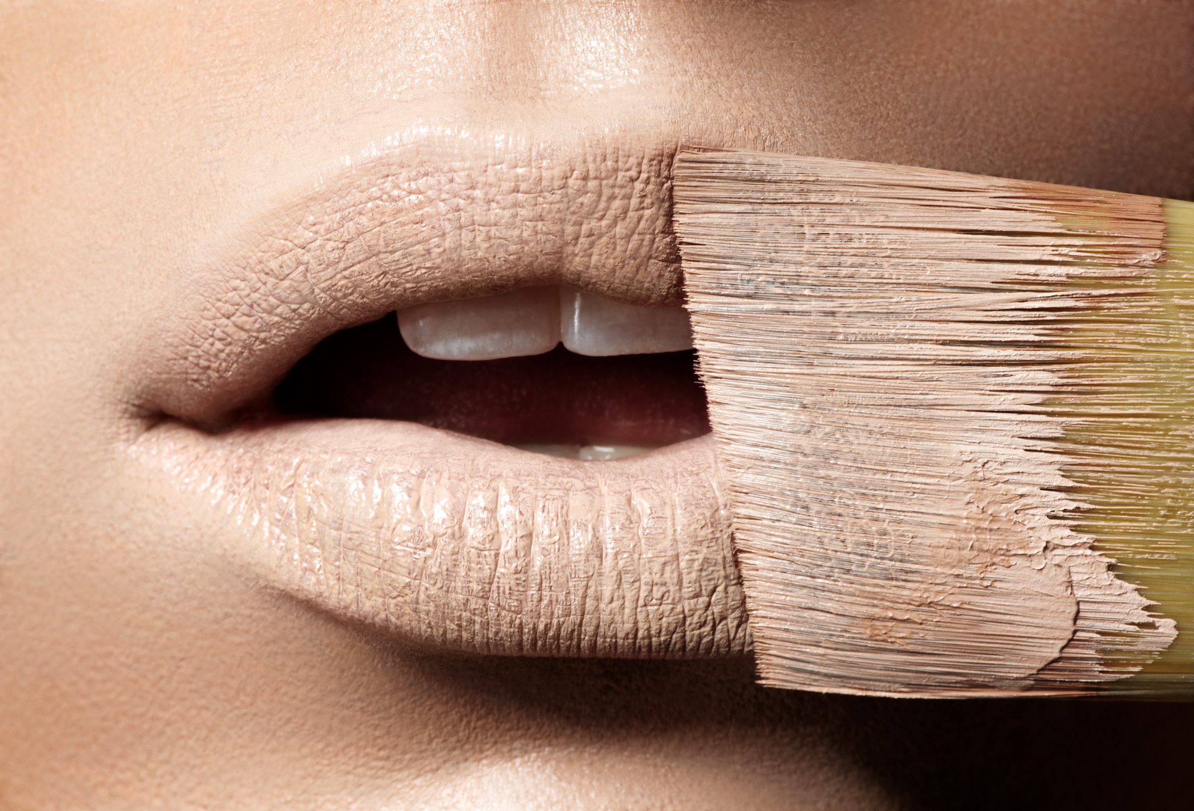 A flat brush drags nude paint across the model's lips in extreme macro.
