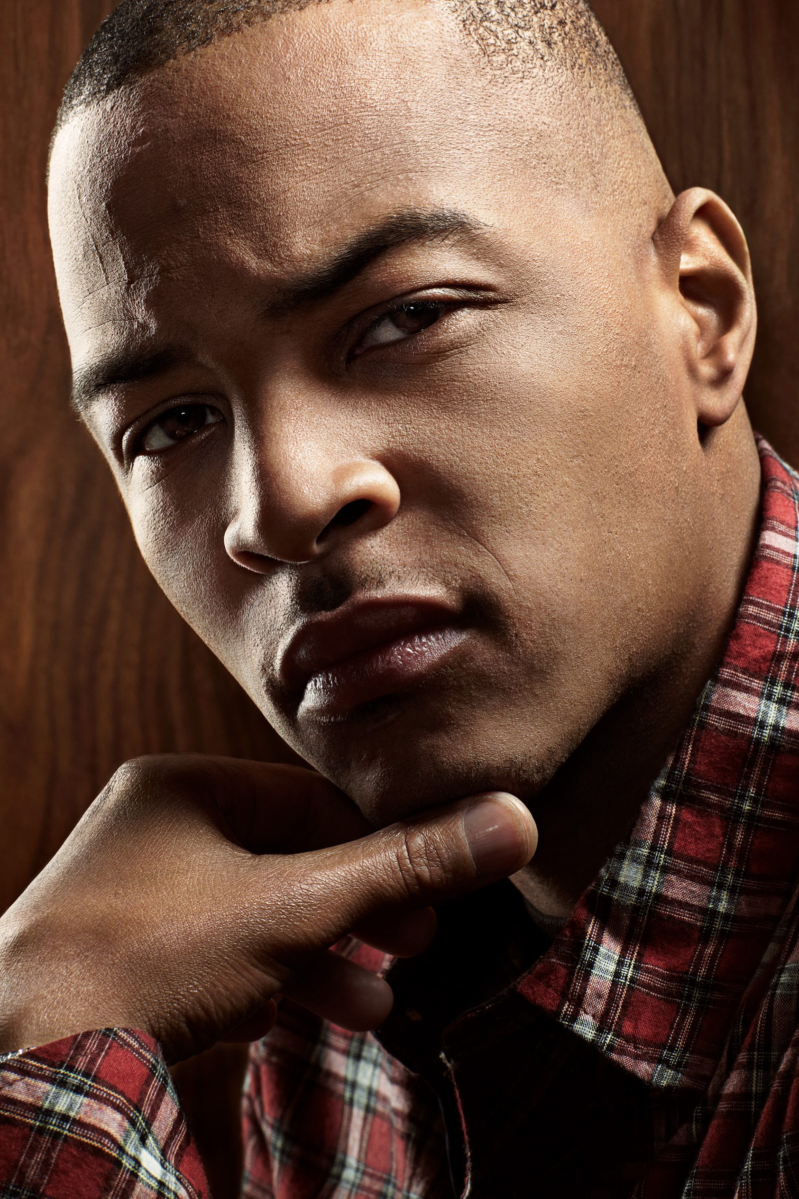 Close-up of T.I. in a red plaid shirt, chin resting on his hand against warm wood paneling.