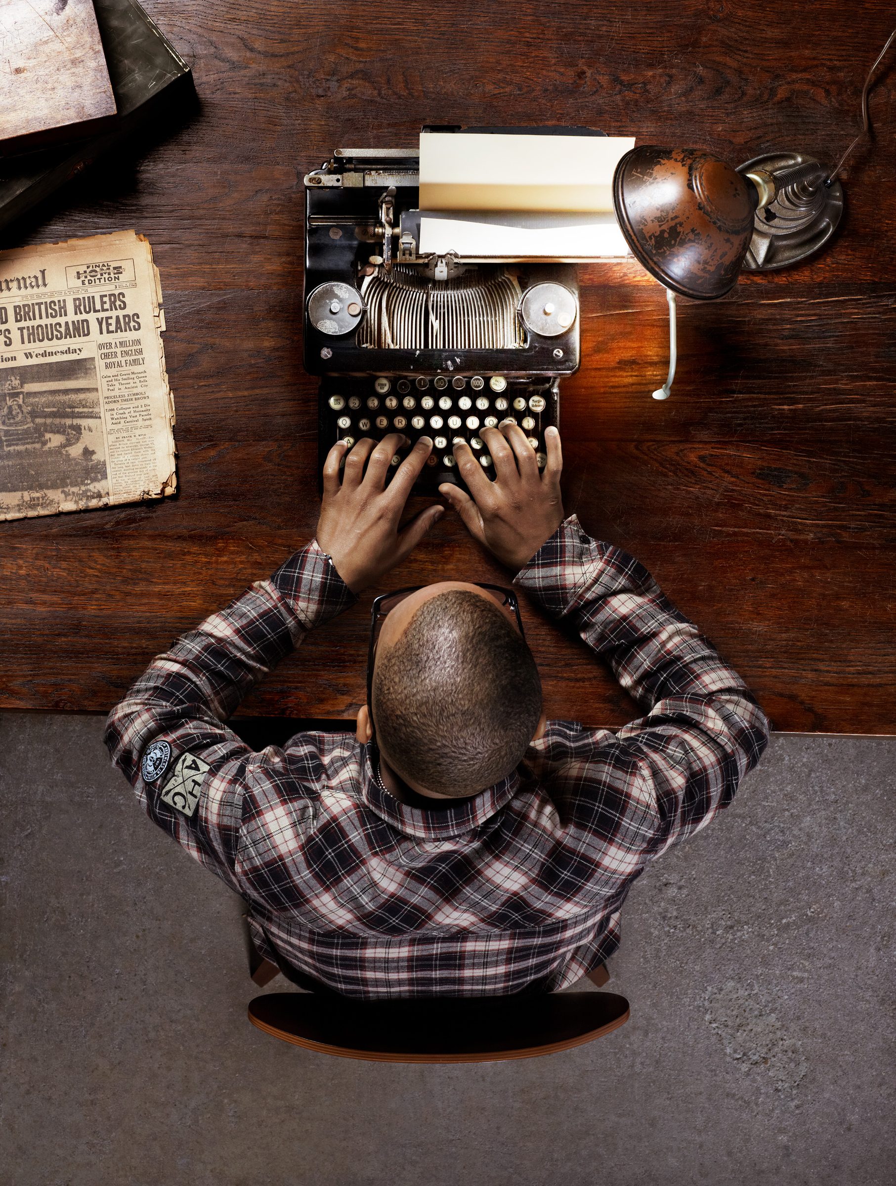 Overhead view of T.I. at a wooden desk, typing on a vintage typewriter beside old newspapers.