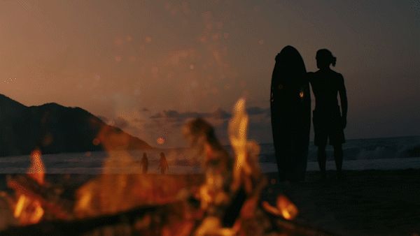 Silhouetted surfers watch the twilight surf beside a roaring bonfire on a Caribbean beach.