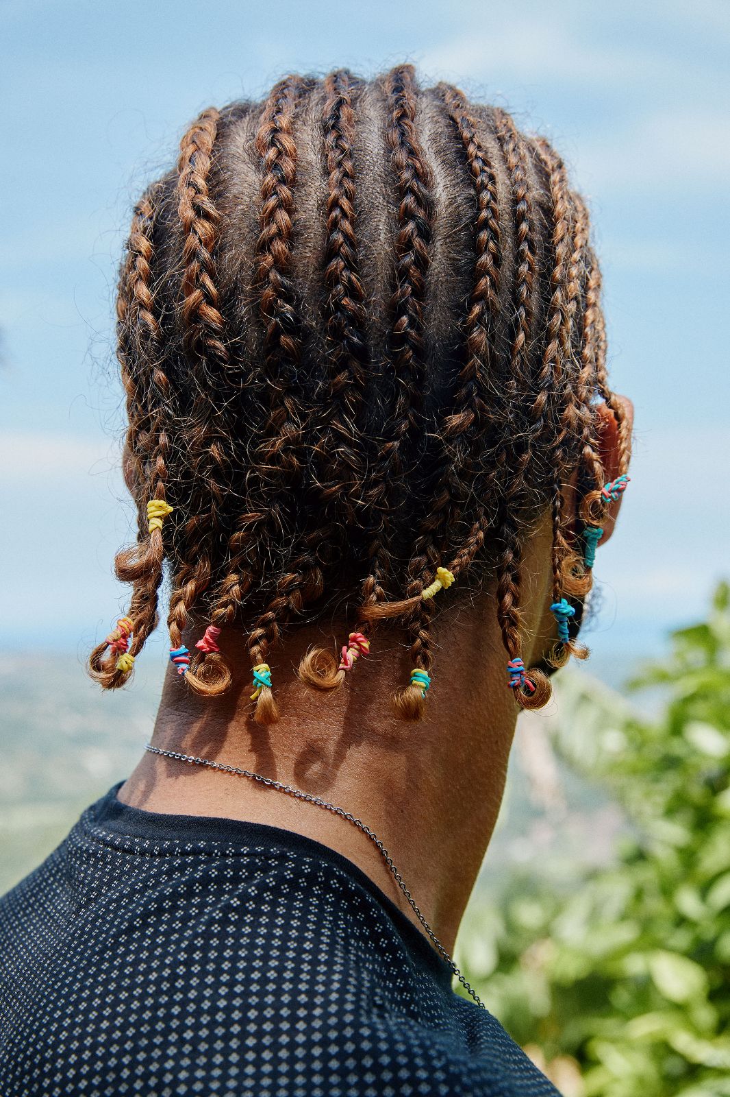 Detail of a man's intricately braided hair with colorful ties facing a coastal landscape.