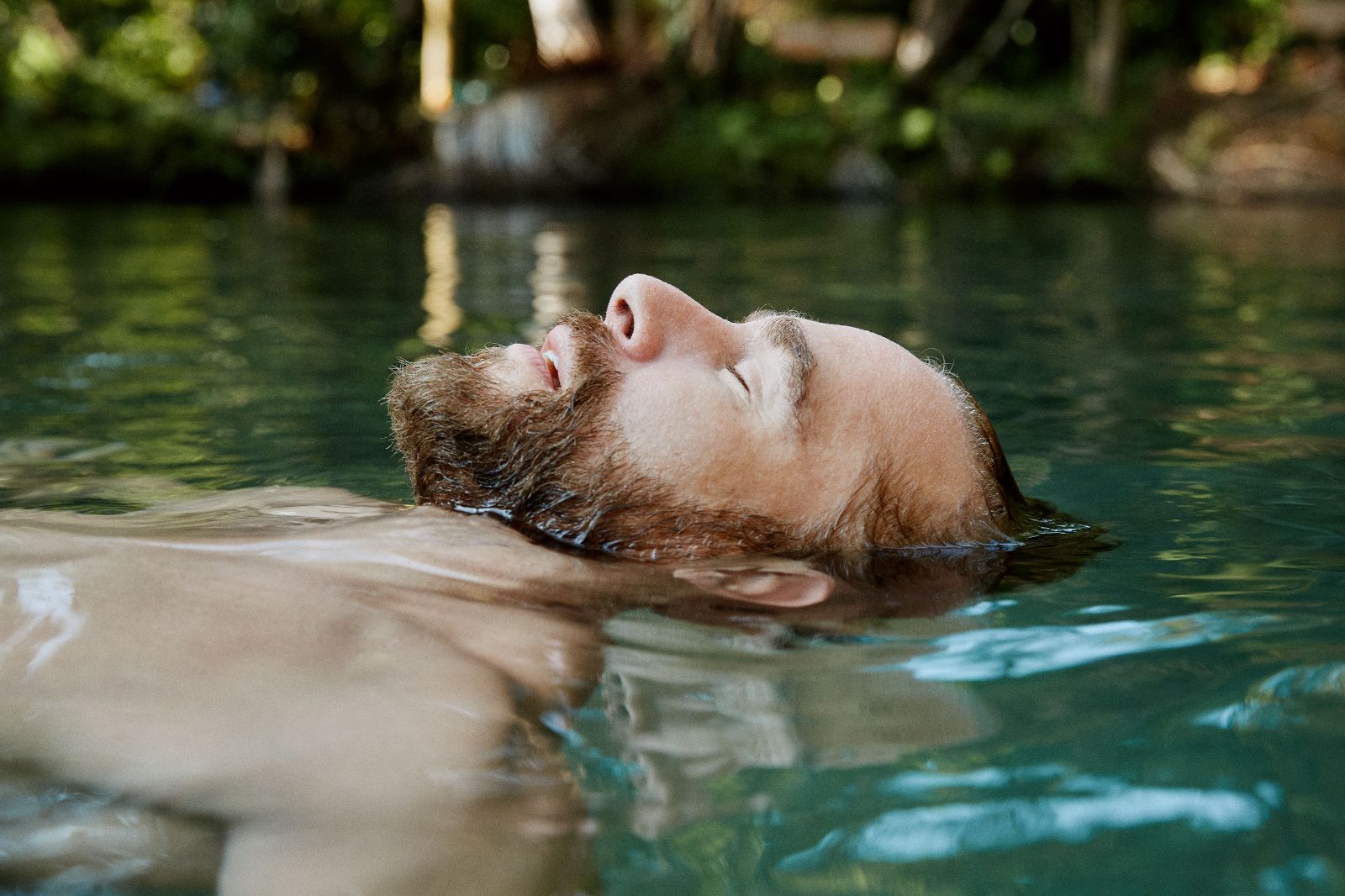 A bearded man floats peacefully on his back in a tranquil, sun-dappled inland cenote.