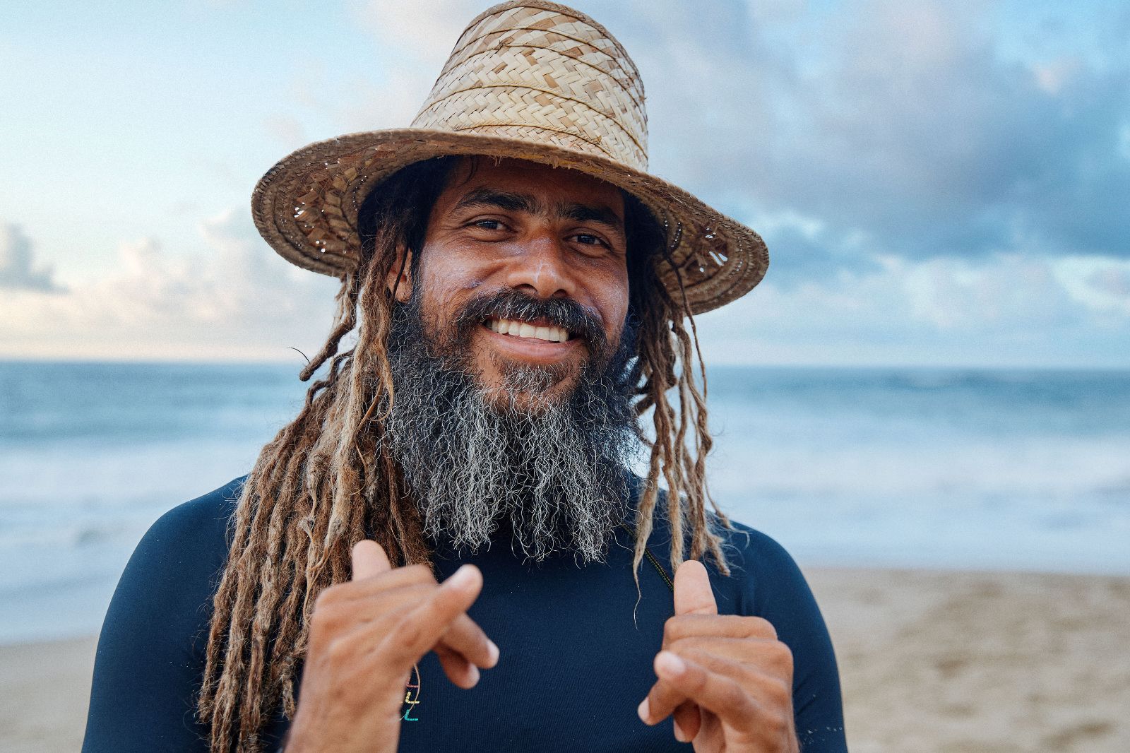 A smiling local man with dreadlocks and a straw hat makes shaka signs on the beach.