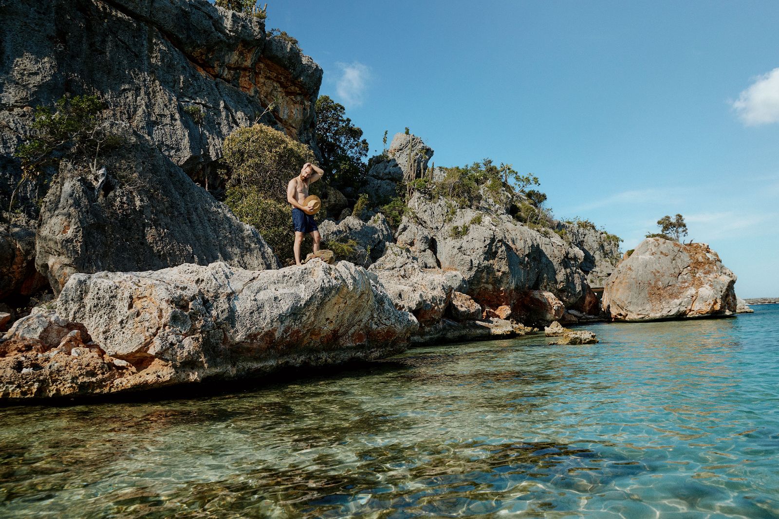 A shirtless man stands on sunlit, rugged limestone cliffs overlooking clear turquoise marine water.