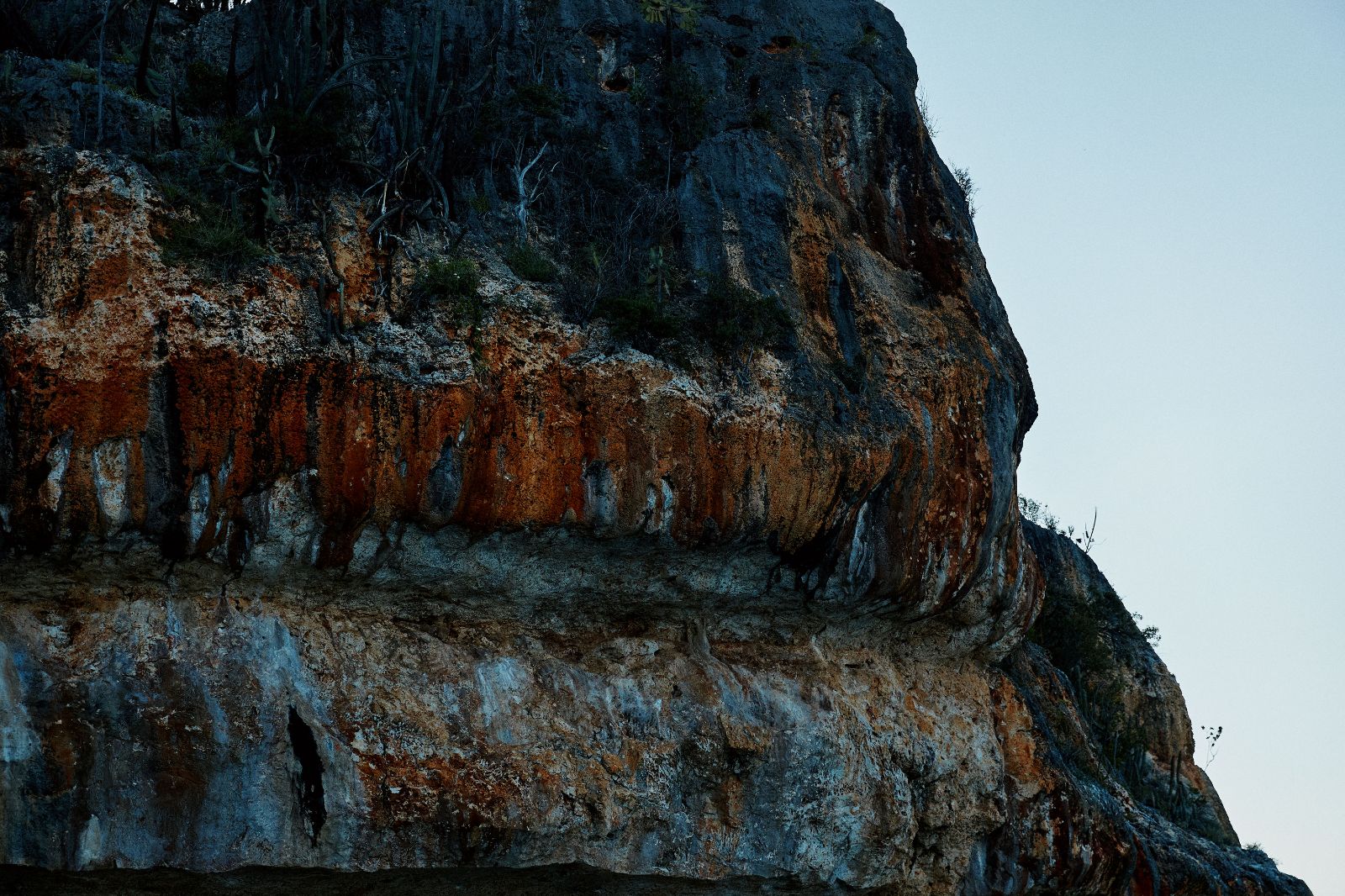 Dramatic rust-colored rock faces of a rugged coastal cliff under a pale blue sky.