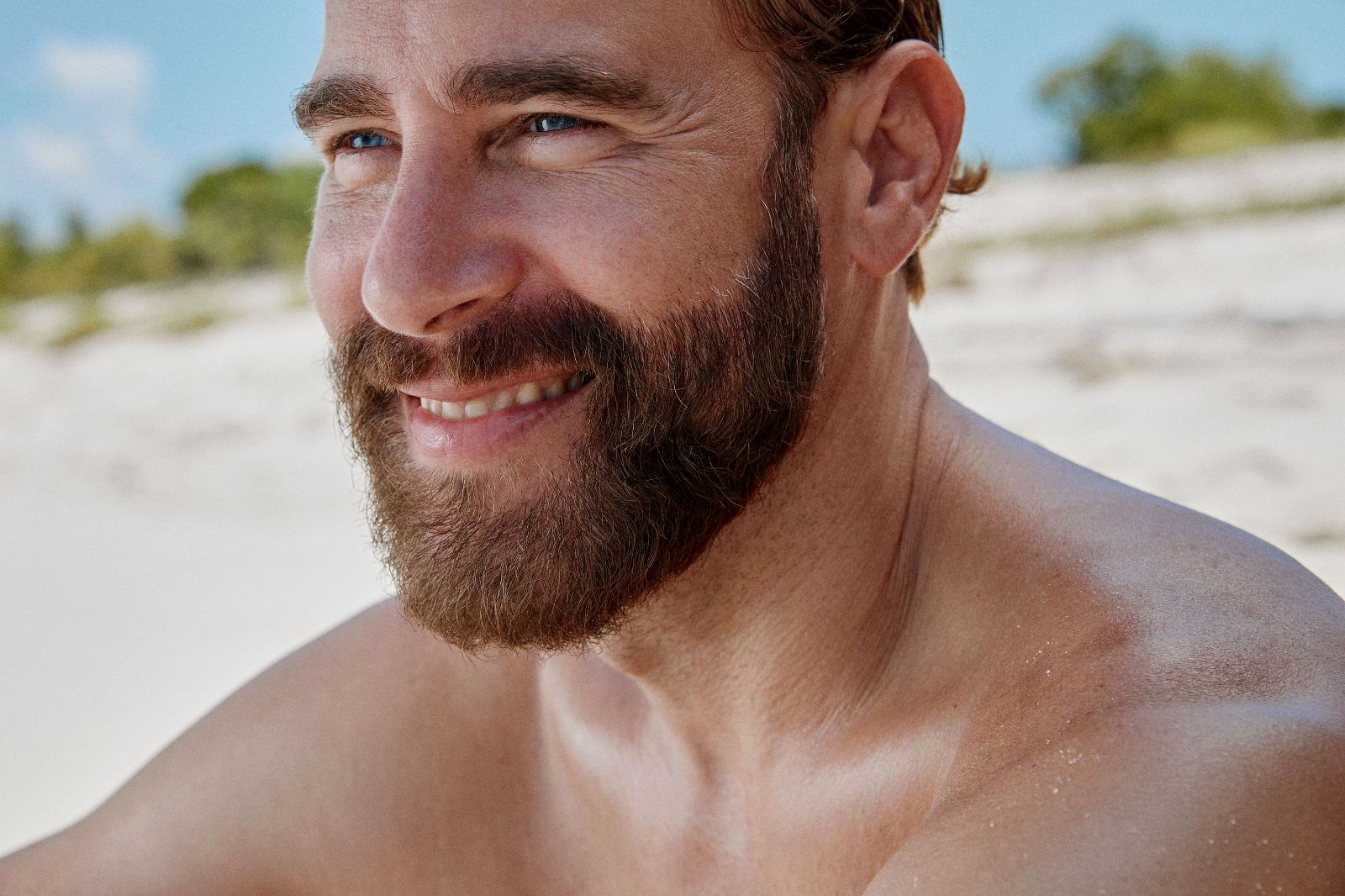 Close-up portrait of a bearded Carlos Delamota smiling warmly on a bright sandy beach.