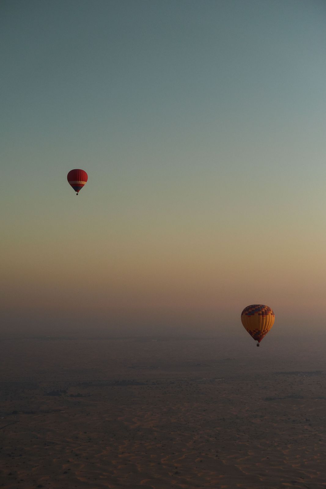 Two hot air balloons drifting peacefully over an expansive, hazy desert landscape.